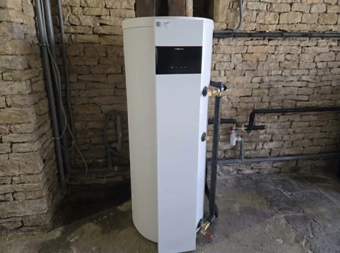 Chauffe-eau thermodynamique blanc ou gris clair installé dans une chaufferie ou buanderie moderne d'une maison résidentielle. L'équipement doit être clairement identifiable comme une pompe à chaleur pour eau chaude sanitaire (technologie thermodynamique avec évaporateur visible ou design caractéristique), installé contre un mur clair dans un intérieur fonctionnel et bien éclairé, montrant la qualité et la performance de la solution.